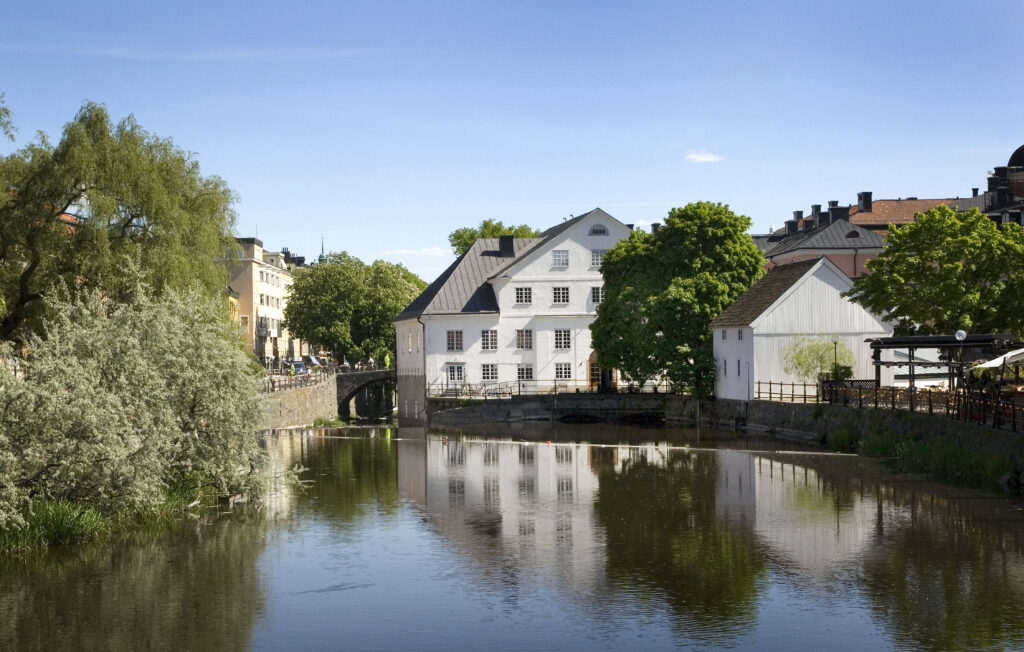 Stor vit byggnad i fyra våningar och med brutet tak i vinkel ligger precis vid vattnet och speglas vackert i sjön.