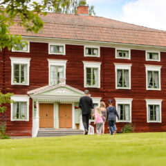 Några personer går över gräsmattan mot en stor rödmålad äldre byggnad med en elegant dekorerad brokvist. Huset är byggt i trä och har tre synliga våningar med fönster med vita karmar.
