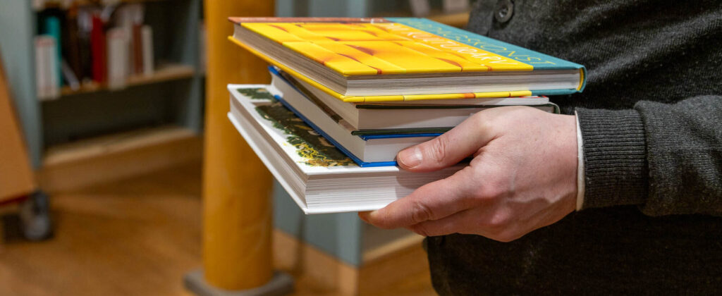 Böcker på hög som bärs av en person. Endast ena handen syns och bokhögen.
