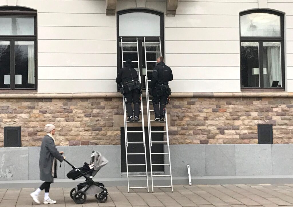 Två hanverkare står på varsin stege som lutar mot fasaden på en äldre byggnad i stadsmiljö. De håller på med att renovera ett fönster. På gatan nedanför passerar en ung kvinna med ett litet barn i en barnvagn.
