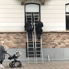 Två hanverkare står på varsin stege som lutar mot fasaden på en äldre byggnad i stadsmiljö. De håller på med att renovera ett fönster. På gatan nedanför passerar en ung kvinna med ett litet barn i en barnvagn.