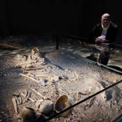 Mörk bild på en grav med skelett i museimiljö och en man som står vid museimontern