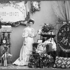 Kabinettsfotografi: Fotografen Mathilda Ranch, sannolikt i sin ateljé, bland blommor och ett blomsterarrangemang. På väggen en skylt med texten "Mathilda Ranch 18 21/5 82 - 19 21/5 07"