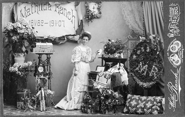 Kabinettsfotografi: Fotografen Mathilda Ranch, sannolikt i sin ateljé, bland blommor och ett blomsterarrangemang. På väggen en skylt med texten "Mathilda Ranch 18 21/5 82 - 19 21/5 07"