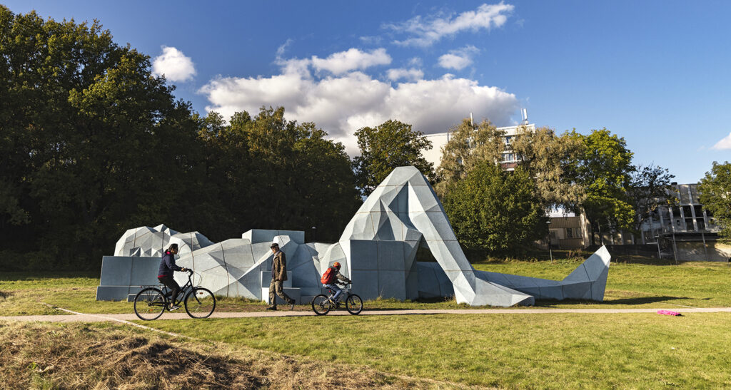 En vit skulptur av en människa i jättestorlek ligger på en gräsmatta framför en gång där tre cyklar åker förbi.