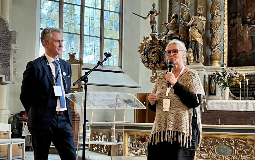 En kvinna med grått uppsatt hår talar i en mikrofon tillsammans med en man med grått hår och kostym. De står framför altaret i en kyrka.