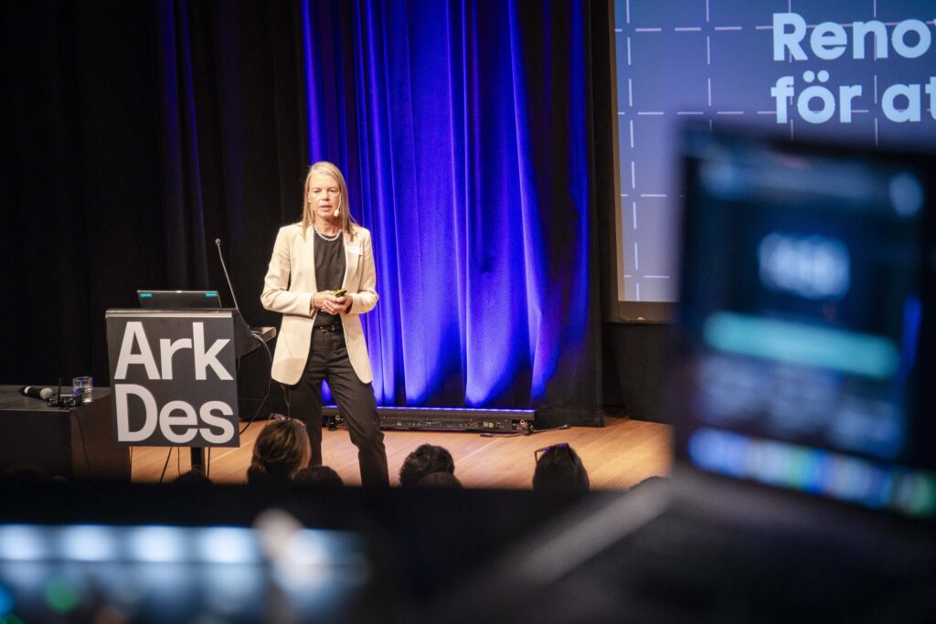 En kvinna står på en scen i en stor konferenssal och talar inför en publik.