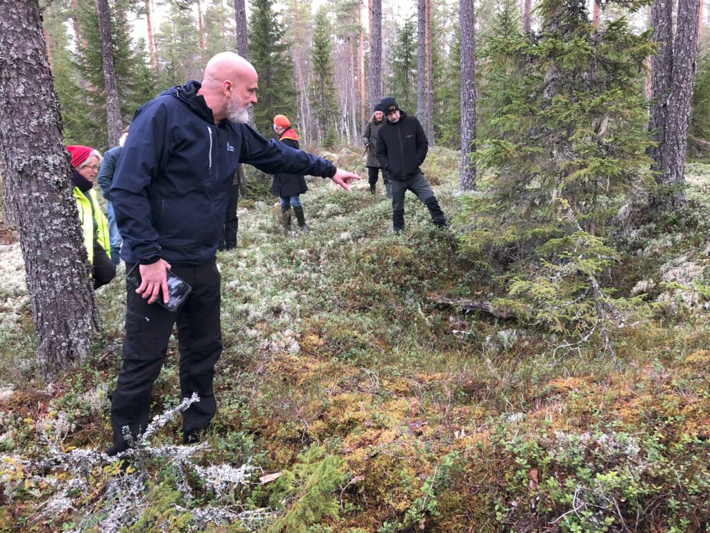 En man står och pekar ner i en grop i skogen. I bakgrunden syns fler personer. Det ser ut att vara höst.