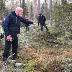 En man står och pekar ner i en grop i skogen. I bakgrunden syns fler personer. Det ser ut att vara höst.