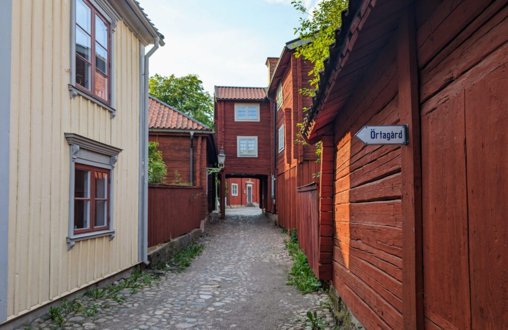 Kullerstensgata omgiven av gamla röda och gula träkåkar. På väggen en skylt om Örtagård.