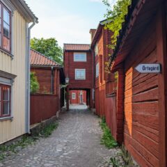 Kullerstensgata omgiven av gamla röda och gula träkåkar. På väggen en skylt om Örtagård.