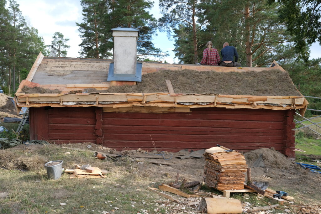 Liten röd timrad byggnad med grönt mosstak som inte täcker mer än halva taket. På nocken har två takläggare tagit paus.