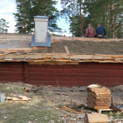 Liten röd timrad byggnad med grönt mosstak som inte täcker mer än halva taket. På nocken har två takläggare tagit paus.